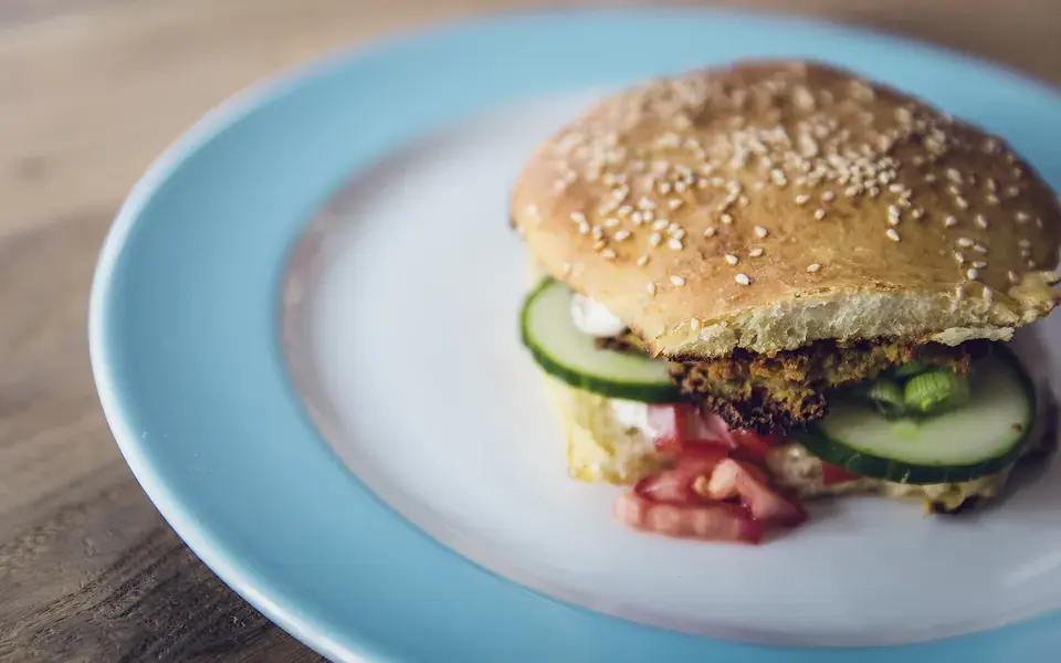 A sesame-seed bun filled with a dark bean patty, cucumber and beetroot, on a pale blue plate.