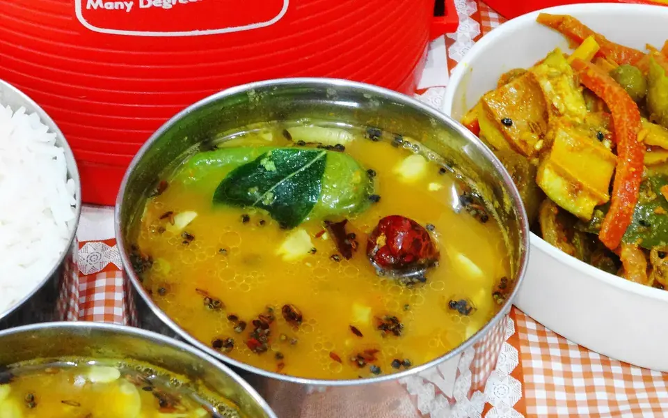 A steel bowl of golden dal scattered with whole spices, beside rice and a vegetable curry.
