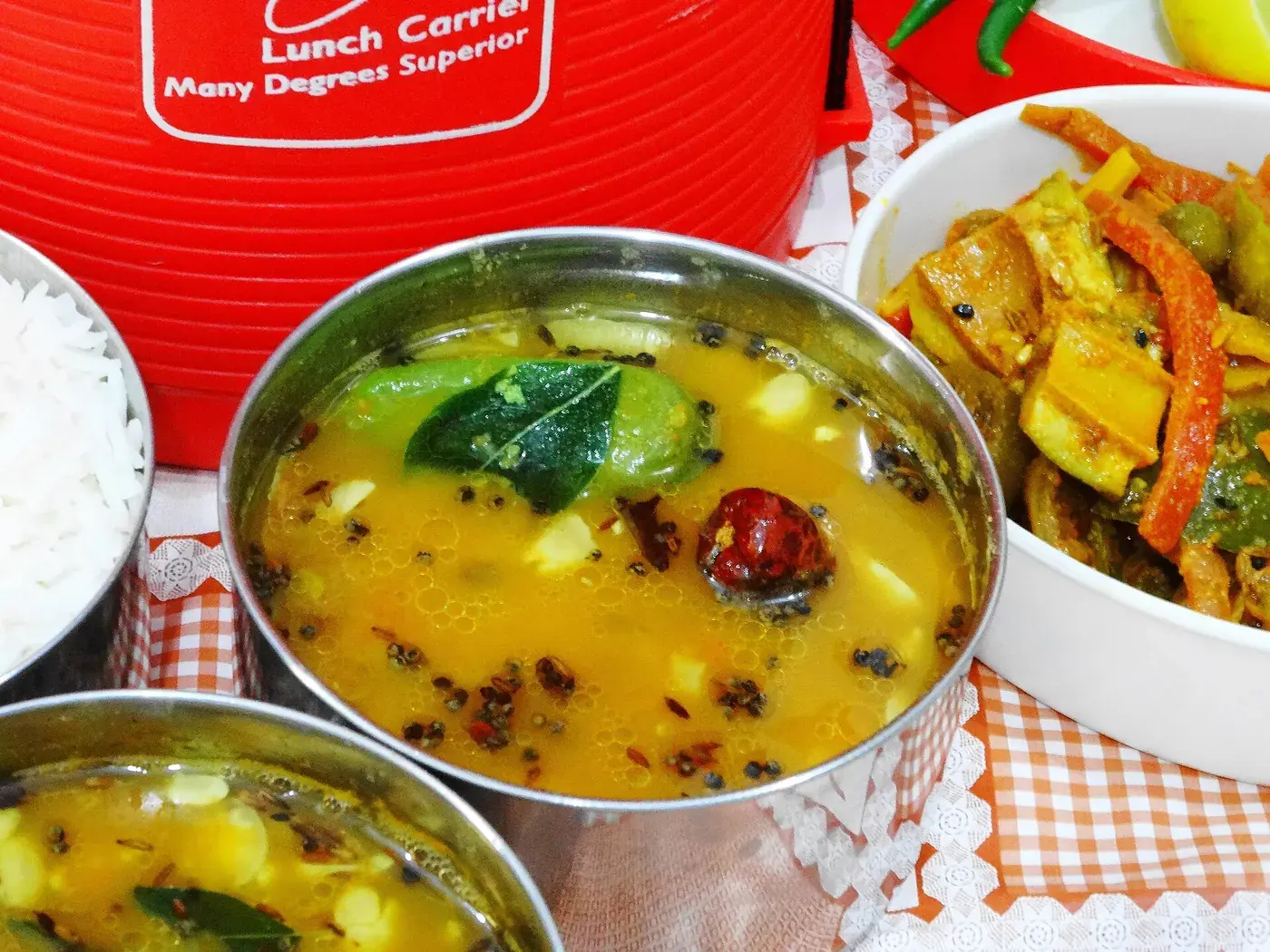 A steel bowl of golden dal scattered with whole spices, beside rice and a vegetable curry.