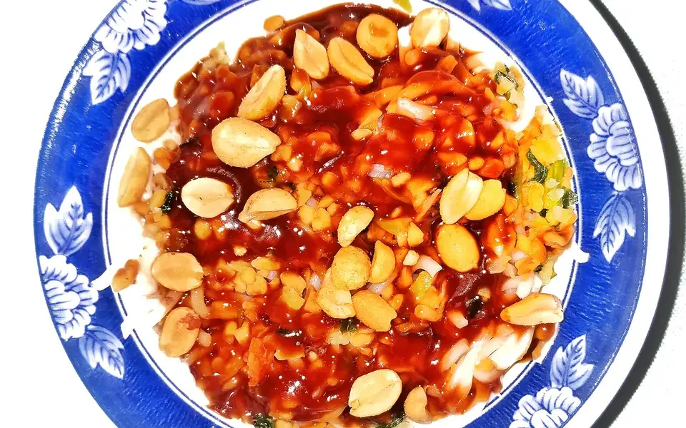 Noodles under a bright chilli and peanut sauce, scattered with whole peanuts, on a blue and white plate.