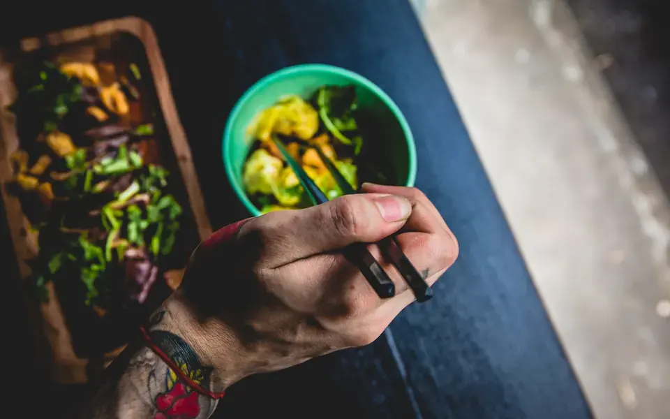 A person holding chopsticks over a bowl of greens, with a board of salad beside it.