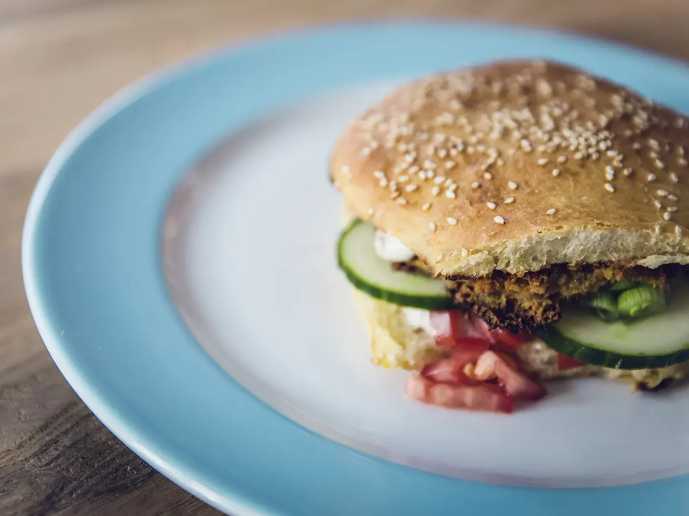 A sesame-seed bun filled with a dark bean patty, cucumber and beetroot, on a pale blue plate.