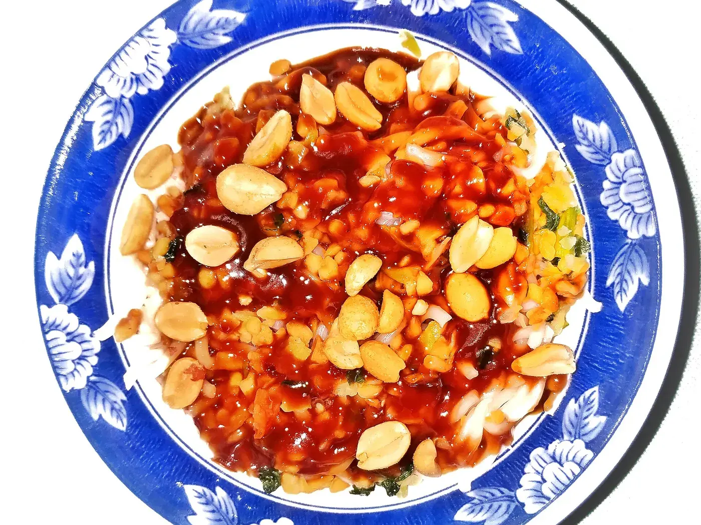 Noodles under a bright chilli and peanut sauce, scattered with whole peanuts, on a blue and white plate.