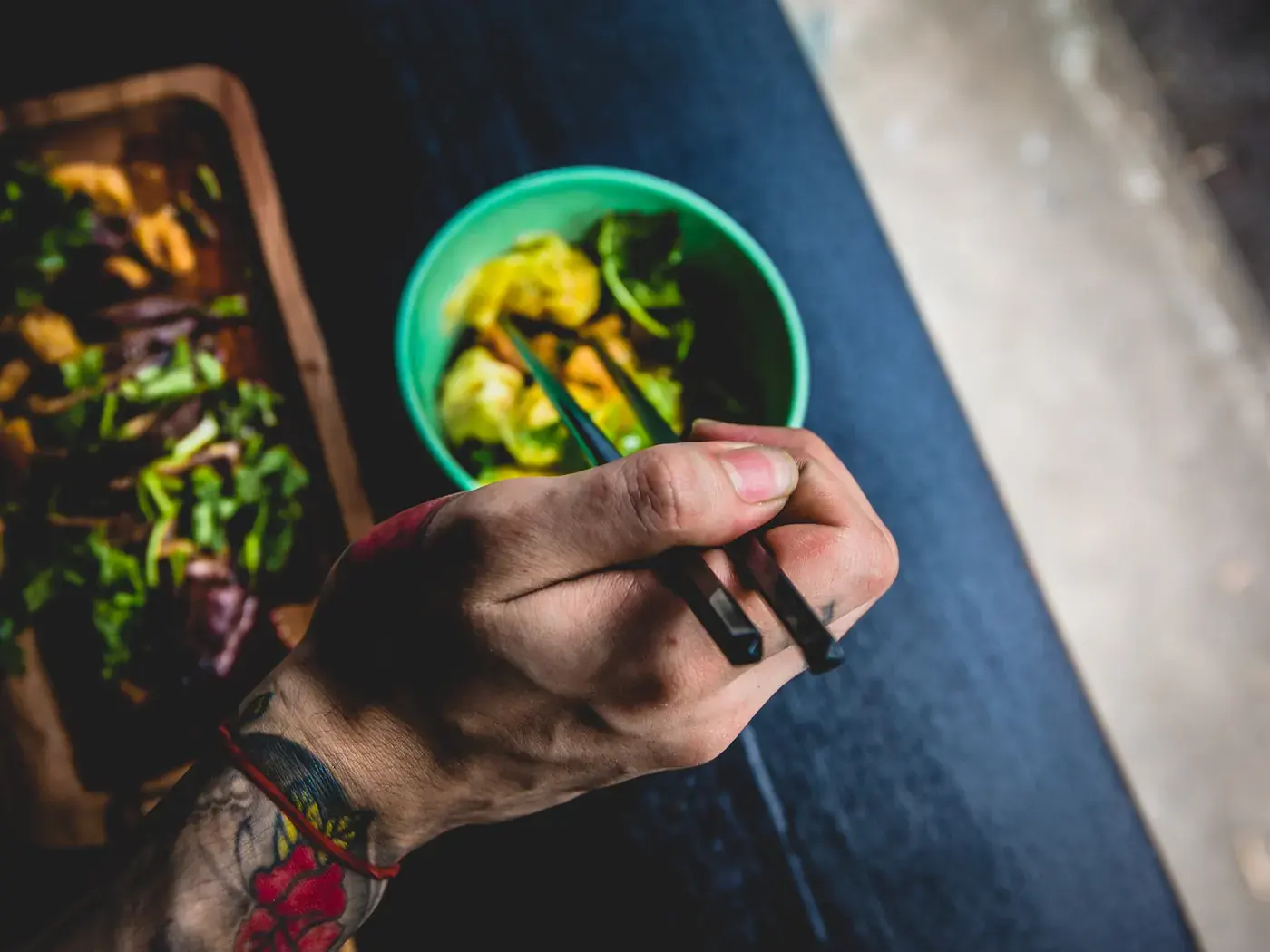 A person holding chopsticks over a bowl of greens, with a board of salad beside it.